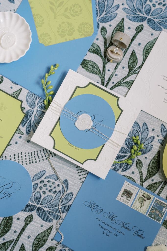 blue and green wedding stationery atop a green floral linen with wedding ring