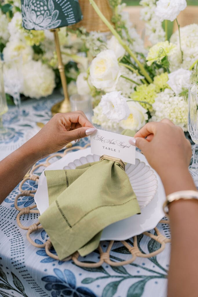 Wedding planner placing place card on top of napkin ring with sage napkin and rattan charger